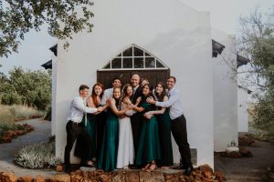 A group of smiling bridesmaids and groomsmen in green dresses and white attire hug outside a rustic chapel.